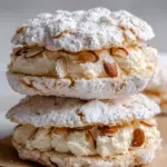 Close-up of two stacked almond dacquoise meringue sandwiches filled with whipped vanilla cream and topped with powdered sugar and toasted almond slices on parchment paper.