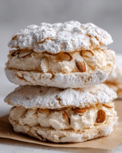 Close-up of two stacked almond dacquoise meringue sandwiches filled with whipped vanilla cream and topped with powdered sugar and toasted almond slices on parchment paper.